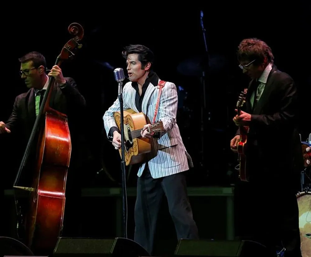Three musicians including a standing bass player a guitarist dressed reminiscent of Elvis Presley and another guitarist perform on a stage