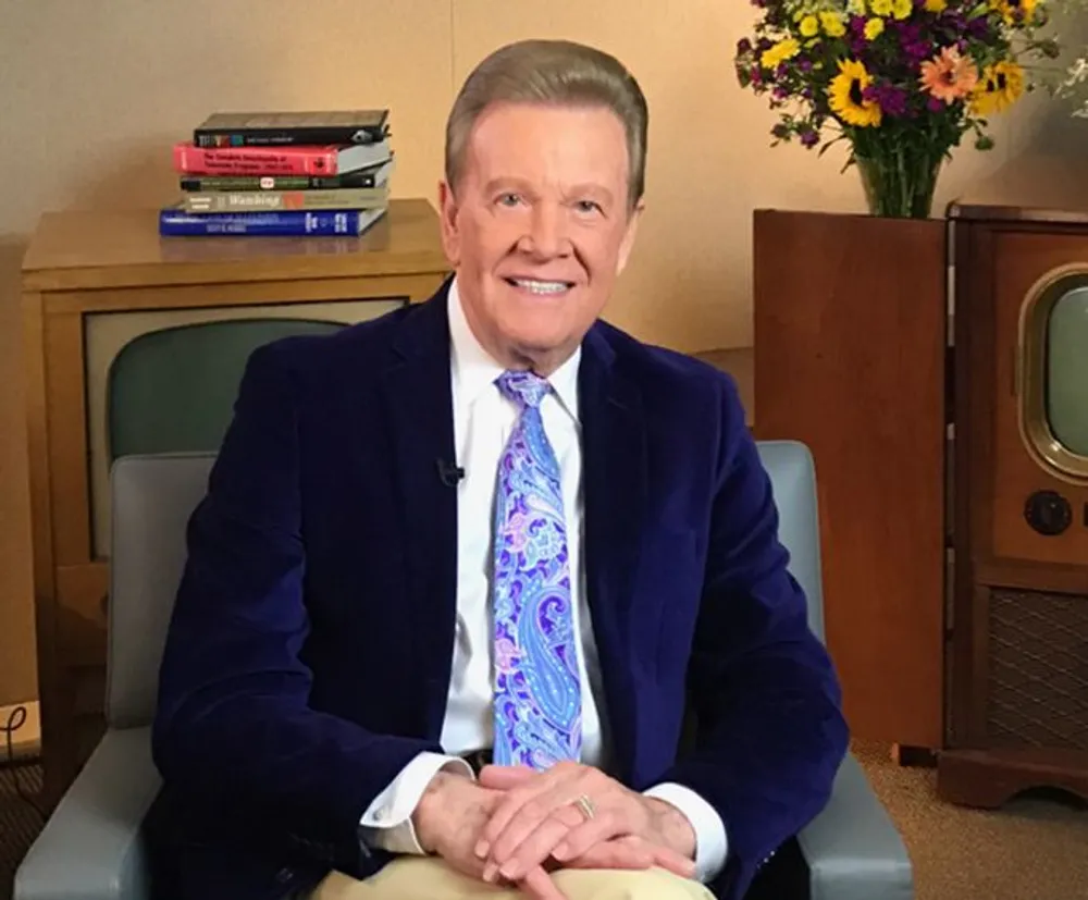A smiling person is seated with their hands clasped over their knees wearing a blue velvet jacket and paisley tie with books and a vintage television set in the background