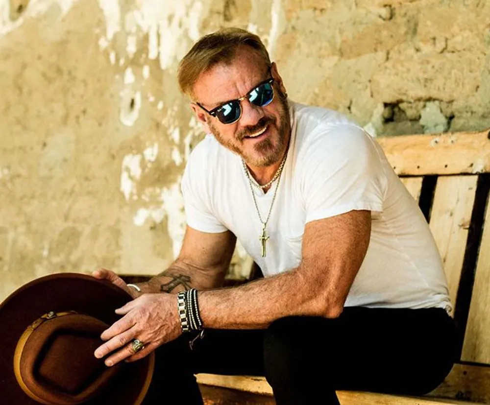 A man with a beard is smiling while holding a brown hat wearing reflective sunglasses and sitting on a wooden bench against a rustic wall backdrop