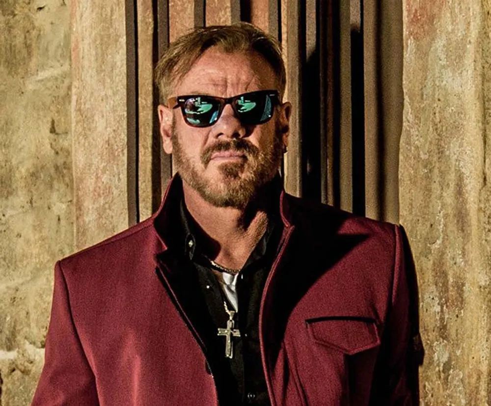 A man with a beard wearing sunglasses and a red jacket stands against a textured backdrop exuding confidence