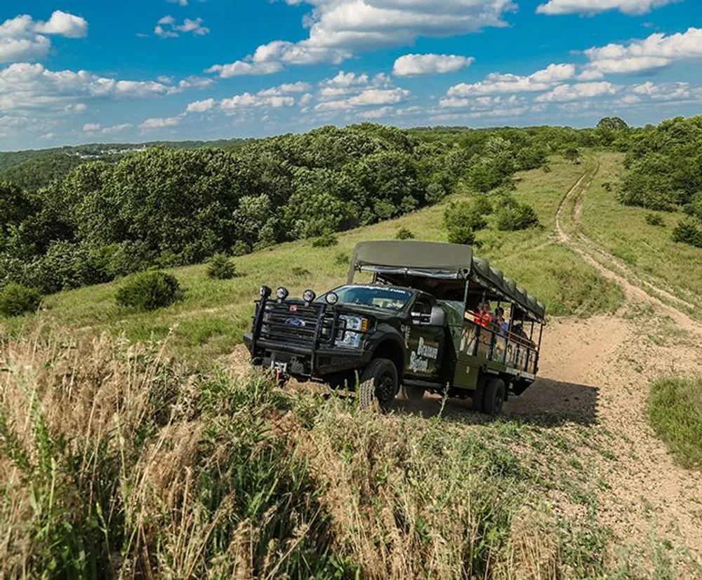 A safari-style vehicle is traversing a rugged trail through a green hilly landscape under a blue sky dotted with clouds