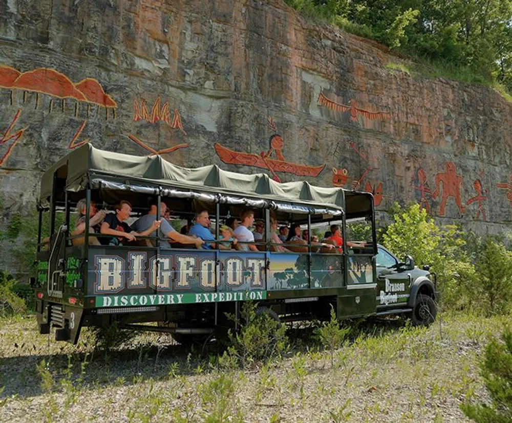 A group of tourists is riding in a vehicle labeled Bigfoot Discovery Expedition passing by a rock face with pictograph-like drawings