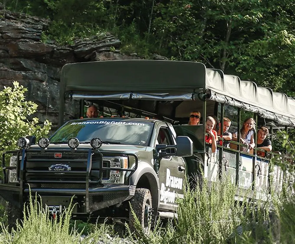 A group of people is on an outdoor expedition in a large open-sided safari-style vehicle driving through a scenic natural environment