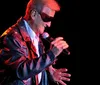 A person wearing sunglasses and a leather jacket is singing into a microphone on stage
