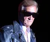 A person wearing sunglasses and a leather jacket is singing into a microphone on stage