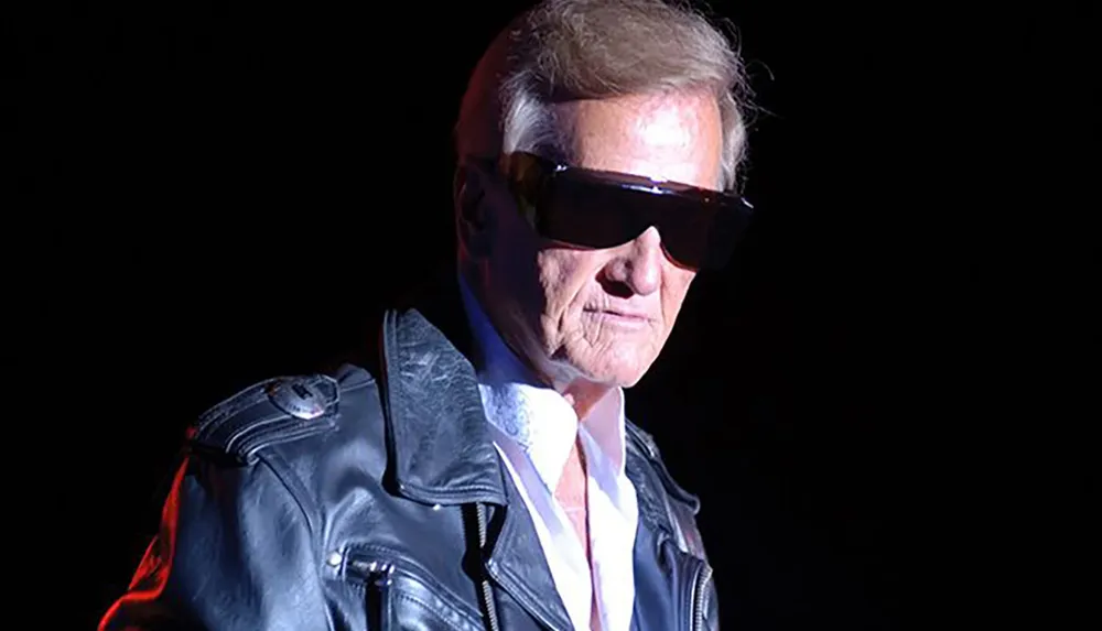 A man wearing dark sunglasses and a leather jacket is looking serious against a dark background