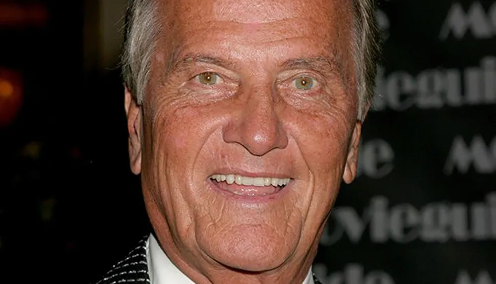 A smiling older man in a suit is posing against a dark background with blurry text