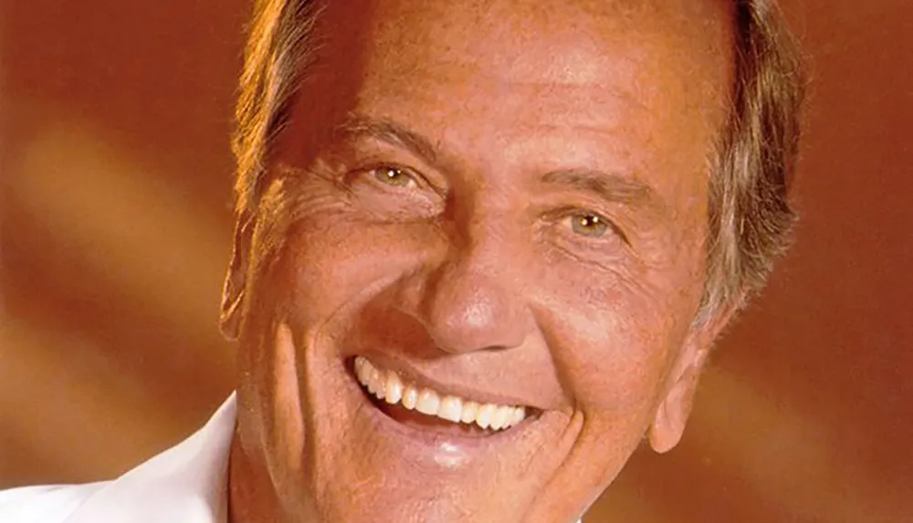 A person with short hair is smiling warmly against a soft warm-hued background