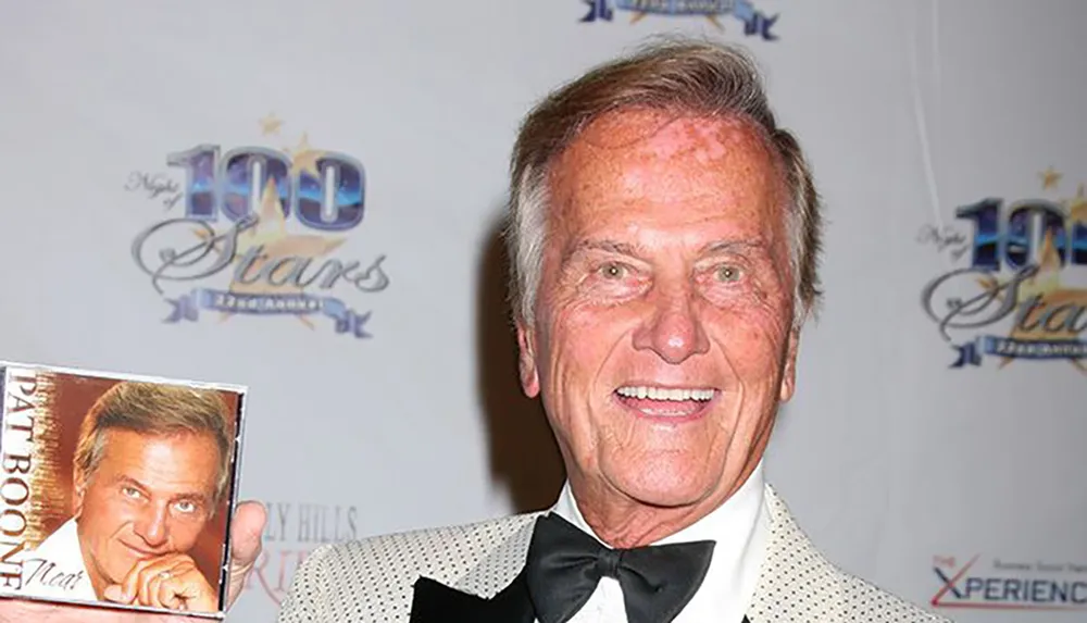 A person is smiling and holding a CD with their picture on it at an event with a Night of 100 Stars backdrop