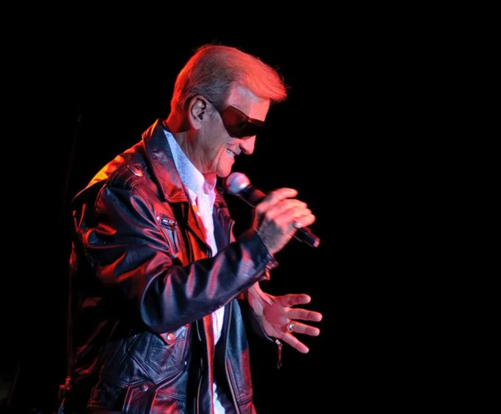 A person wearing sunglasses and a leather jacket is performing with a microphone on a stage illuminated by red lighting