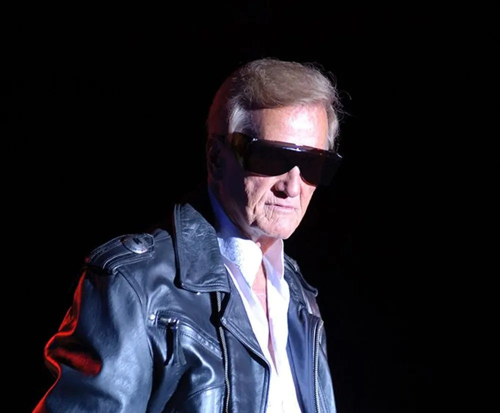 A man is wearing sunglasses and a leather jacket giving him a cool and confident appearance against a dark background