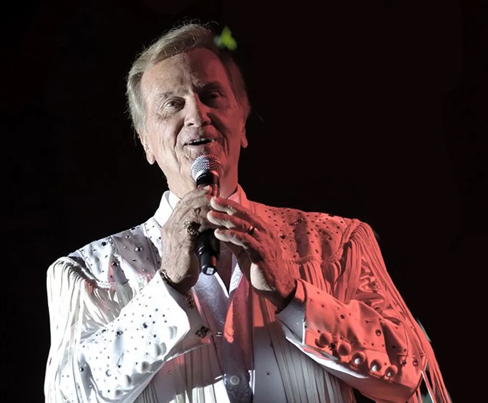 A performer wearing a white rhinestone-decorated outfit is singing into a microphone on stage