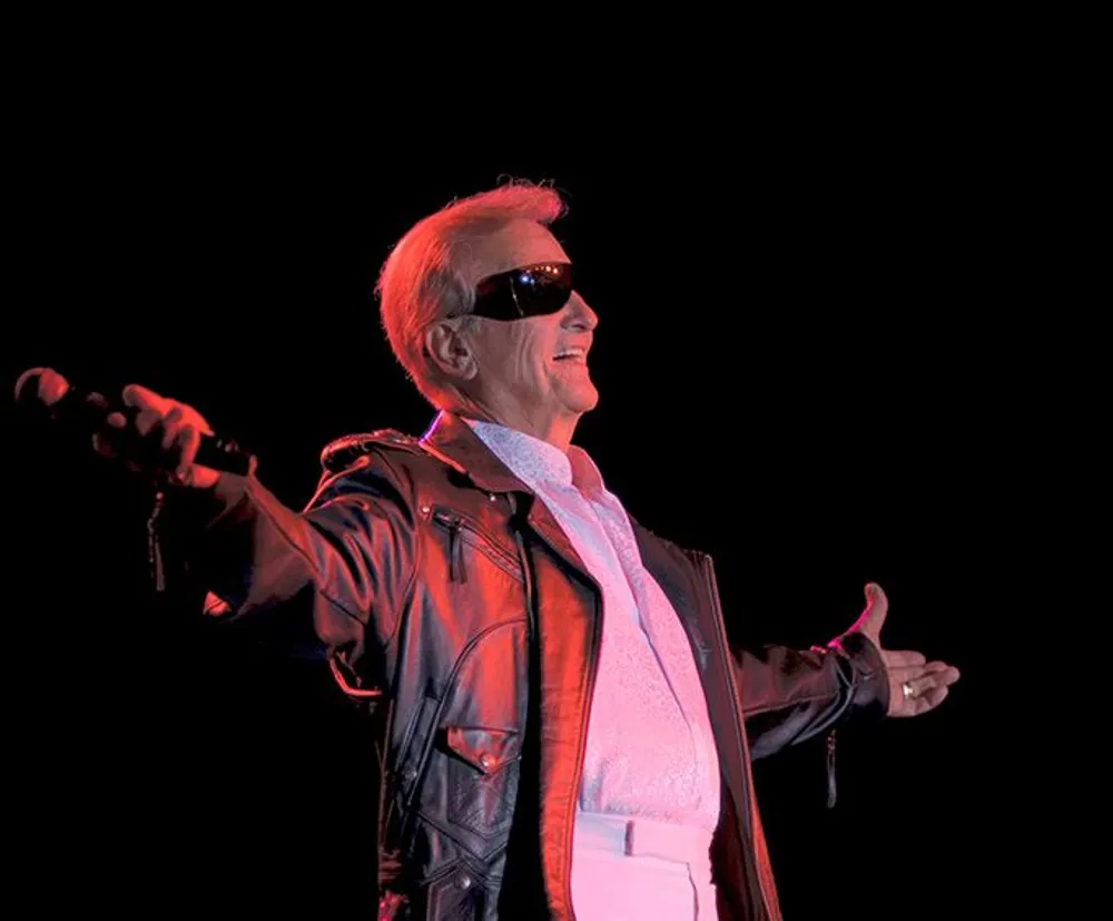 A man in sunglasses and a leather jacket holds a microphone while striking a confident pose on a dark stage with a hint of red lighting