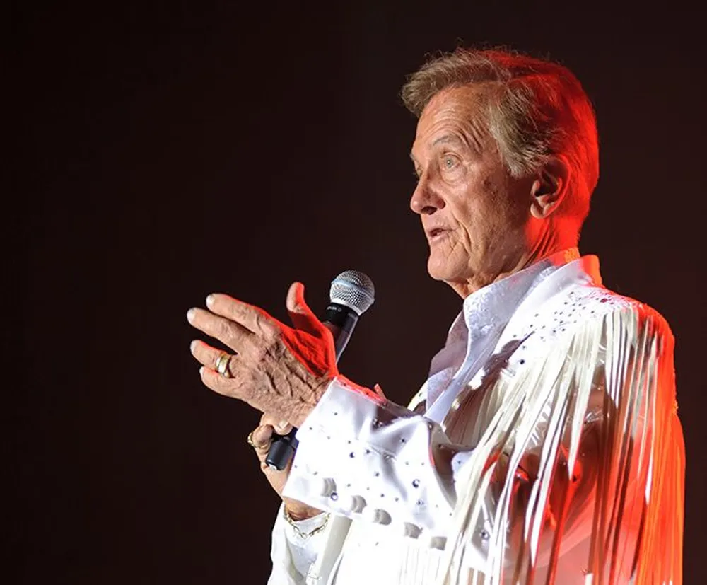 A performer is on stage wearing a white rhinestone-studded outfit speaking into a microphone with a focused expression