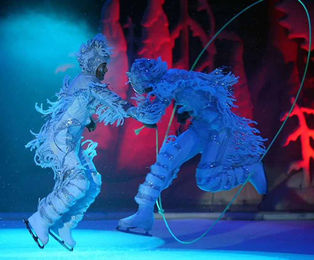 Two performers in ornate blue costumes are ice skating as part of a theatrical show with intricate lighting setting a dramatic backdrop