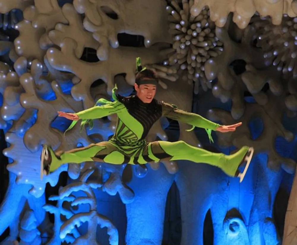 A performer dressed in a vibrant green costume is caught mid-air against a blue and white abstract backdrop giving the impression of flying or leaping