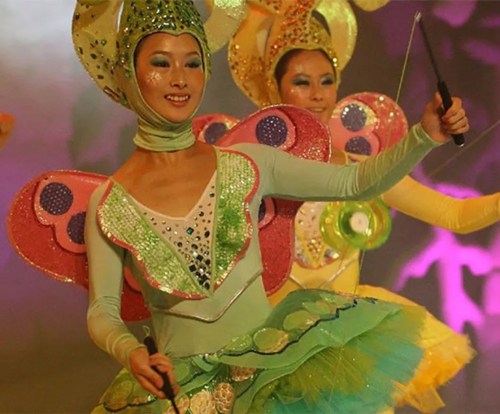 The image shows two performers dressed in colorful butterfly costumes complete with wings and headdresses against a blurred stage backdrop