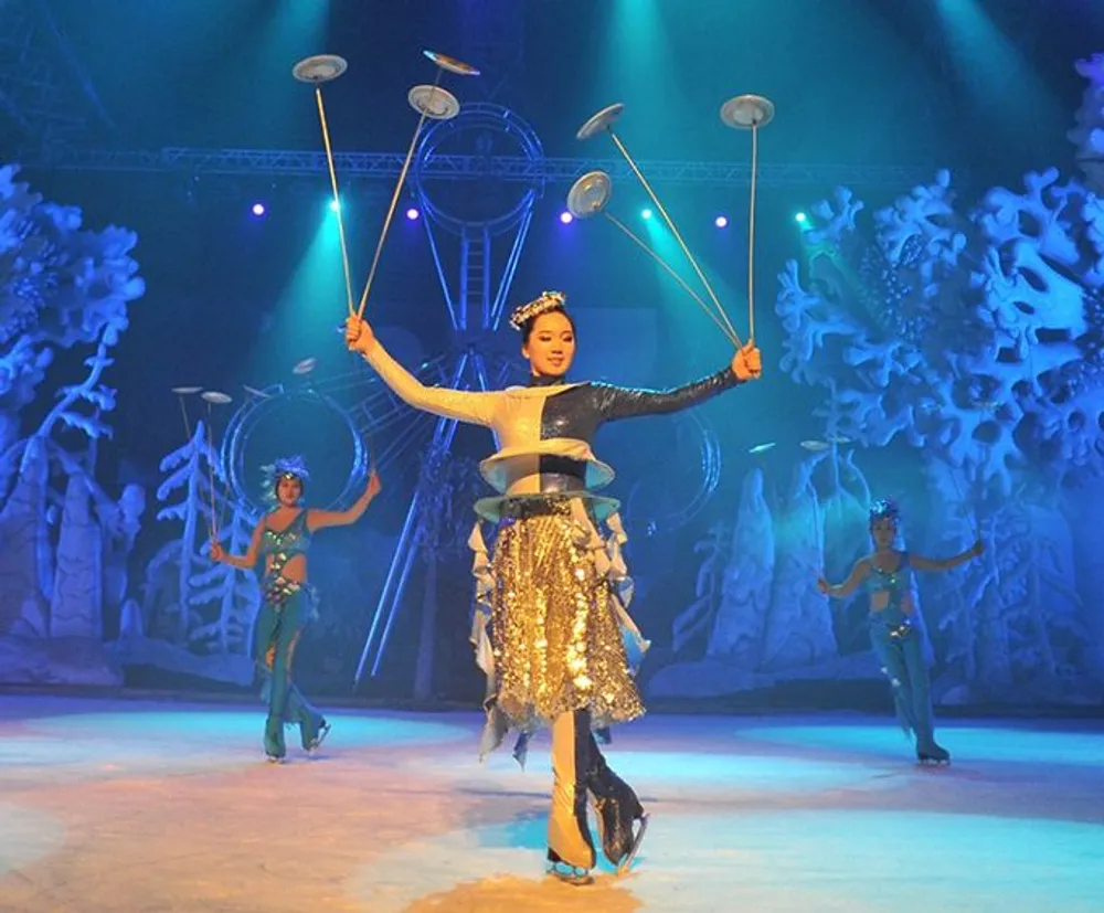 A performer is displaying a plate-spinning act on stage with other artists performing in the background against an icy-blue set design