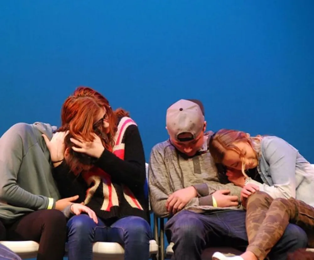 The image shows a group of people comforting each other appearing to be in a state of sadness or grief