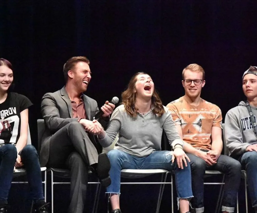 A group of people are sitting on stage sharing a cheerful moment with one man laughing heartily and holding a microphone while a young woman next to him laughs openly surrounded by others who seem amused