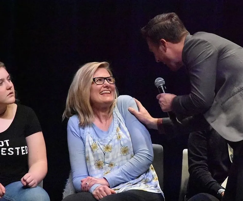 A man holding a microphone is attentively speaking to a smiling woman seated among an audience while another person looks on from the side