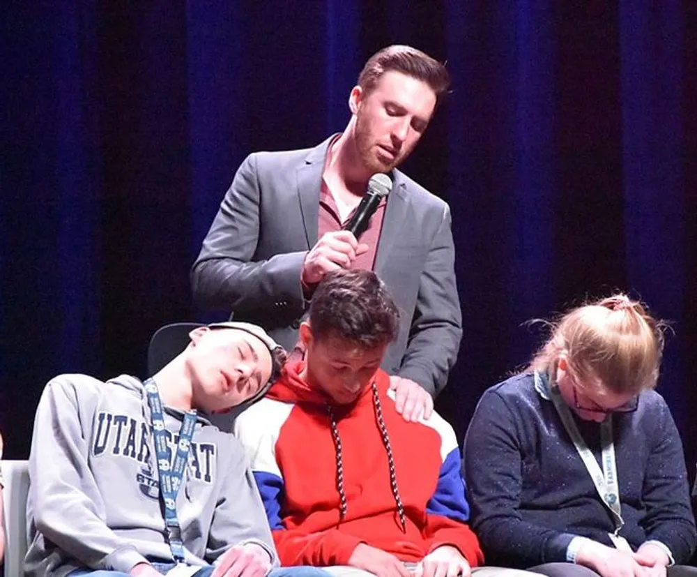 A man with a microphone speaks while three individuals appear to be asleep or in a trance-like state on stage