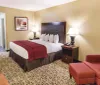A neatly made hotel bed with white linens and a red bedspread in a room with a patterned carpet and neutral decor