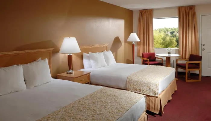 The image shows a neatly arranged hotel room with two beds side tables with lamps a small dining area and a window with curtains