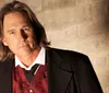 A person with long hair is wearing a formal suit with a red patterned vest standing against a textured wall