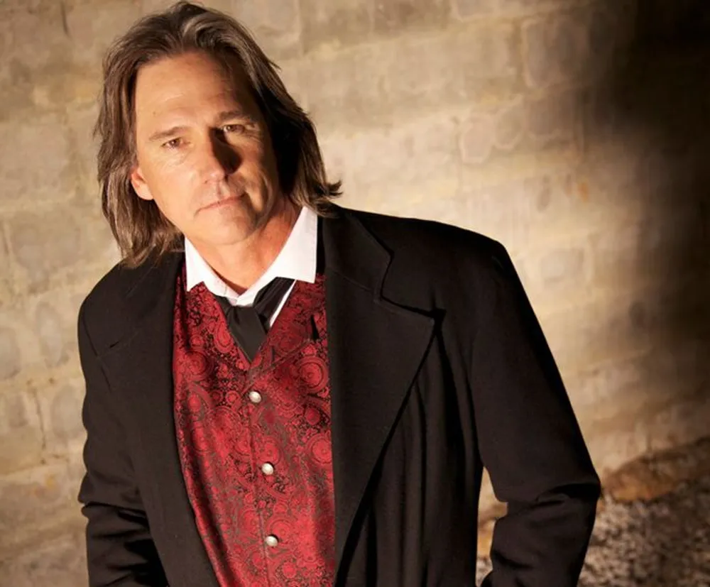 A person with shoulder-length hair is wearing a black suit jacket over a paisley-patterned red vest and a white shirt against a stone-wall background