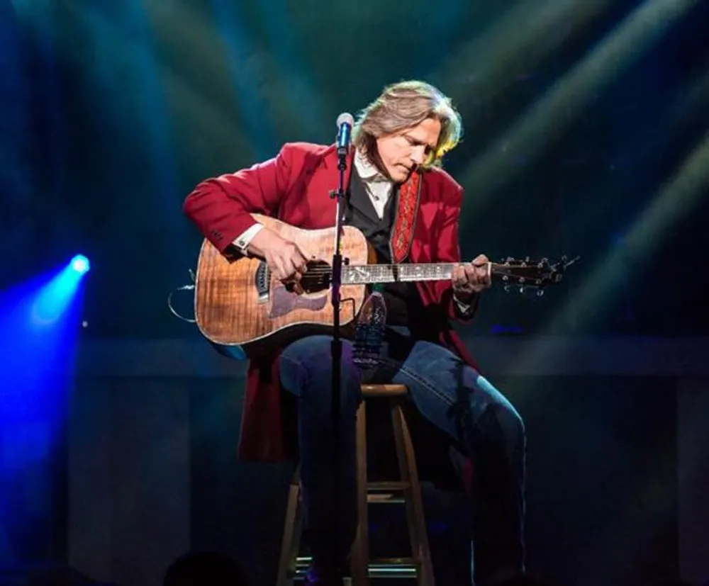 A musician is seated on stage playing an acoustic guitar illuminated by dramatic stage lighting