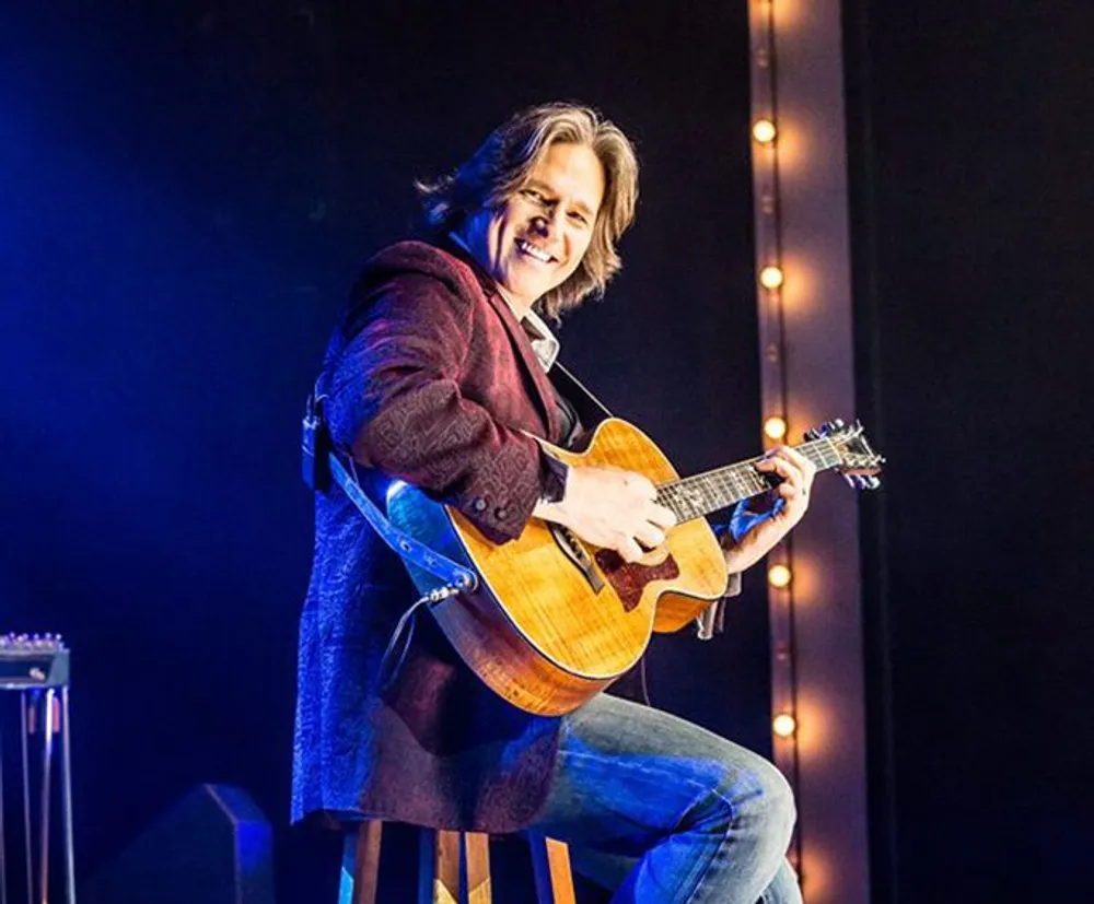 A musician with a warm smile is playing an acoustic guitar while seated on stage