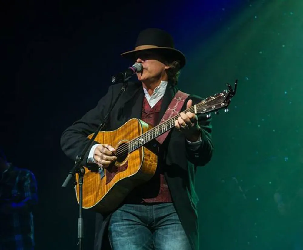 A musician in a hat is playing an acoustic guitar and singing into a microphone on stage illuminated by a spotlight with a blue and green background