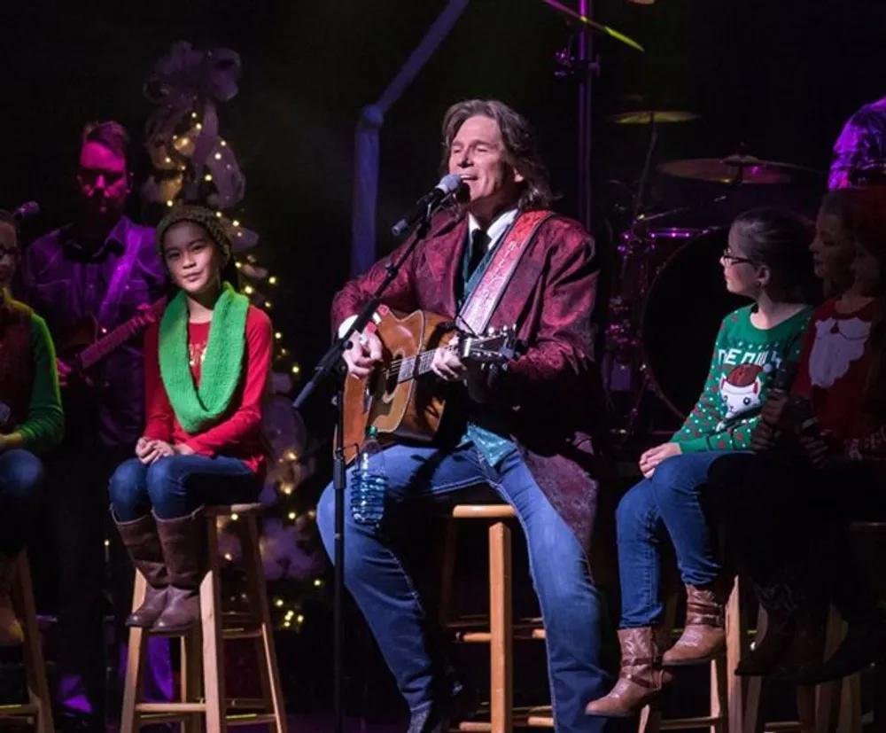 A musician is performing on stage with a guitar while singing into a microphone accompanied by people some of whom appear to be children dressed in festive holiday attire