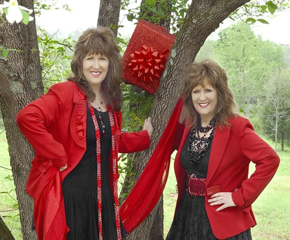 Two women dressed in festive red and black attire are smiling near an outdoor tree adorned with a large red bow