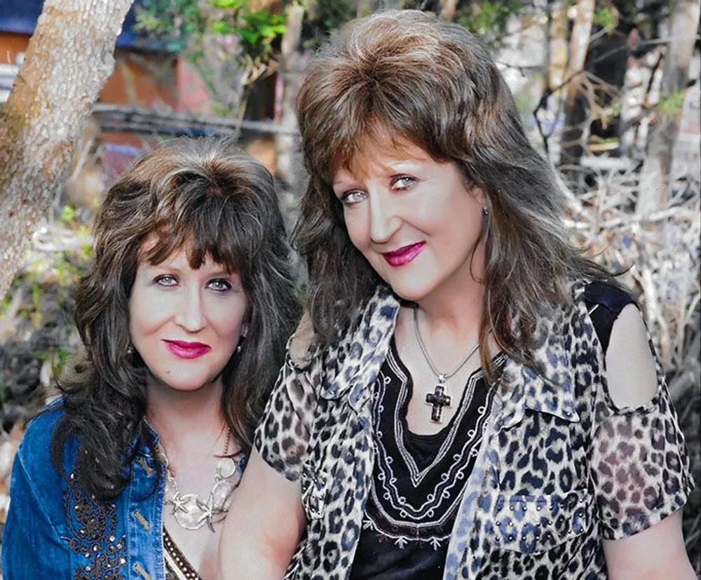 The image shows two people posing closely together both wearing makeup and styled hair with one person in a leopard print top and natural foliage in the background