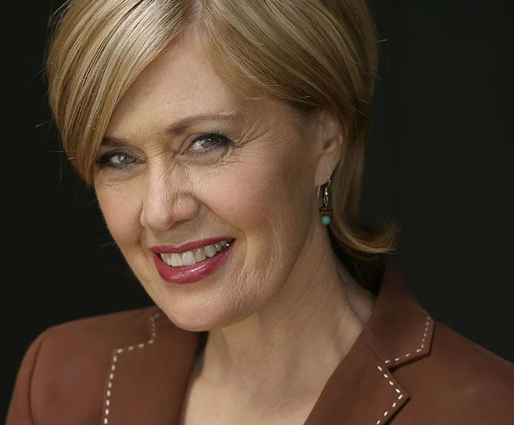 The image shows a smiling woman with blond hair wearing a brown top and turquoise earrings set against a dark background