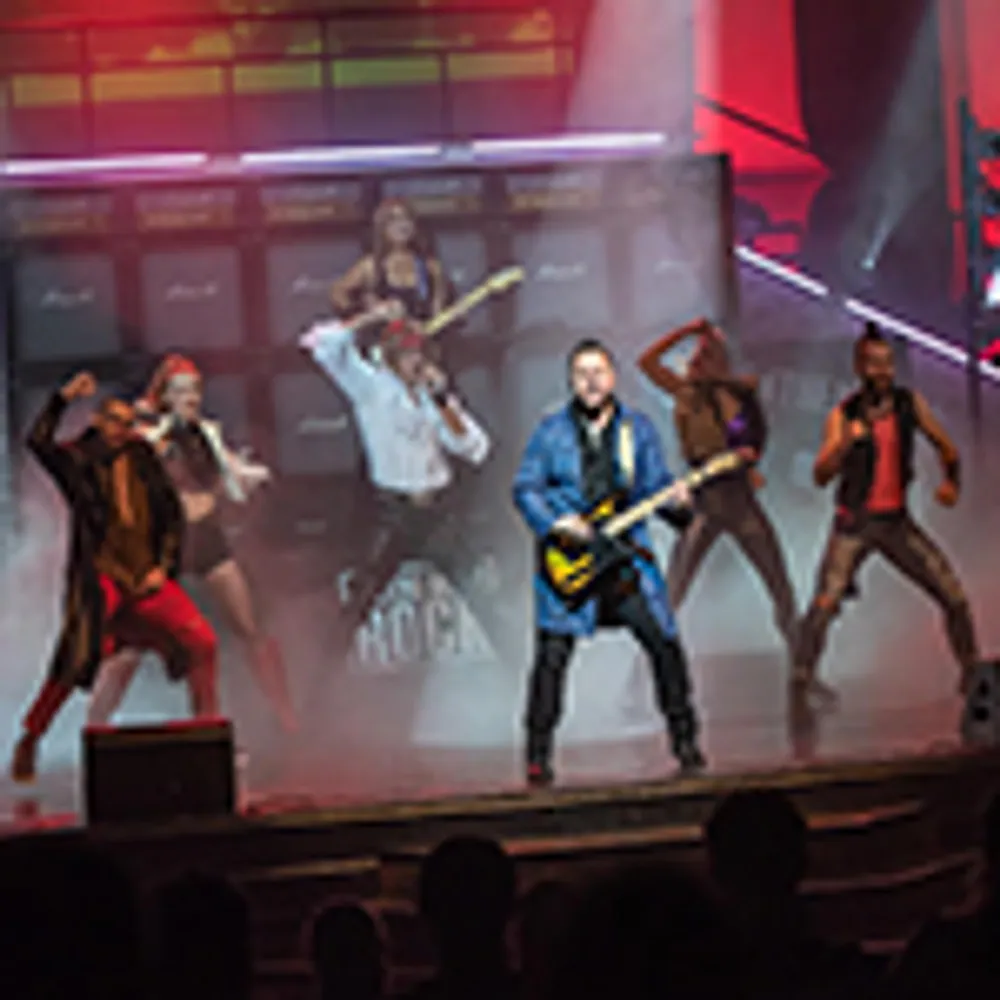 A group of performers energetically play instruments and dance on a stage amid colorful lighting effects