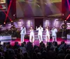 A group of performers in white outfits sings on stage accompanied by musicians while an audience watches