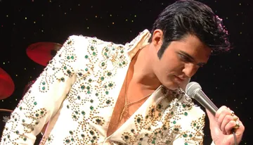 A performer in a bejeweled white suit is singing into a microphone on stage.