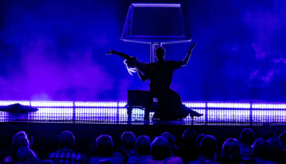 A silhouetted performer dances with a square prop under illuminated blue lights in front of an audience