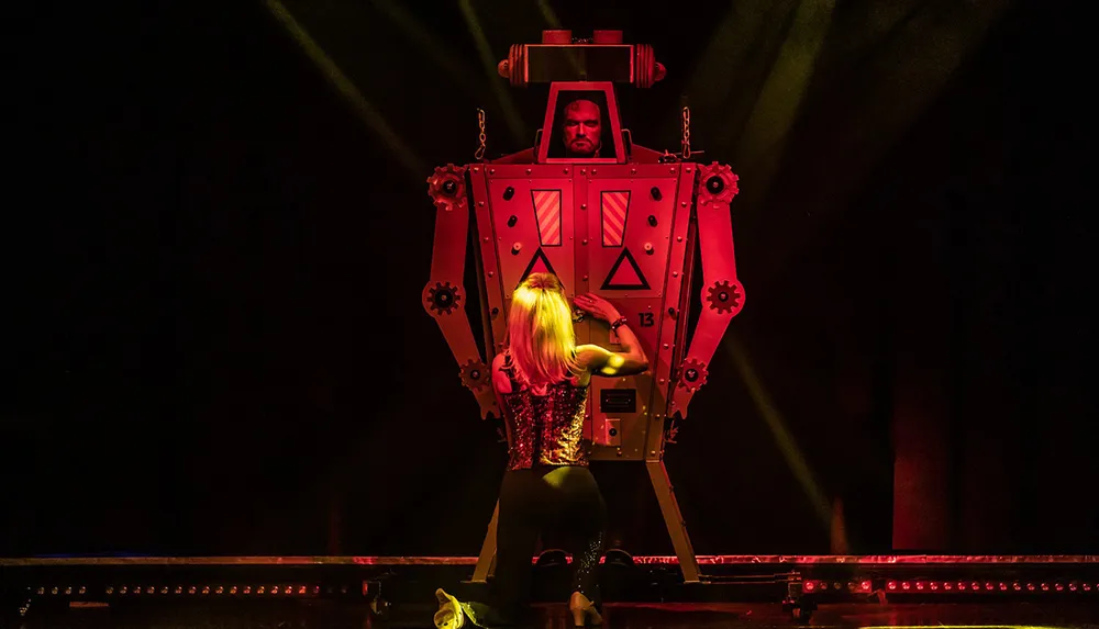 A performer on stage interacts with a large mechanical contraption that features a mans stern face at the top