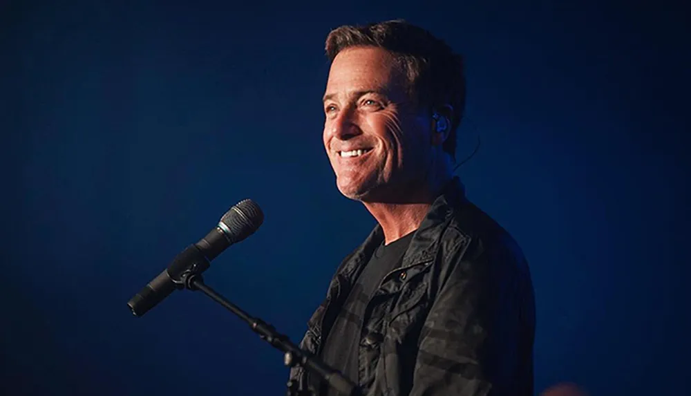 A man with a warm smile stands in front of a microphone on a stage with a dark blue background