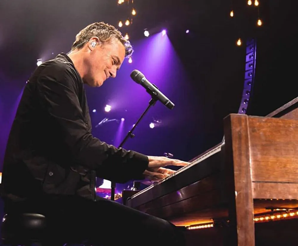 A person is performing at a piano on a stage with a microphone in front and stage lights in the background