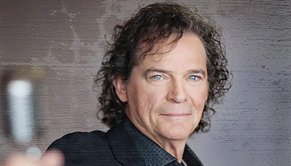 The image shows a confident middle-aged man with curly hair wearing a black blazer posing for the camera against a textured grey background