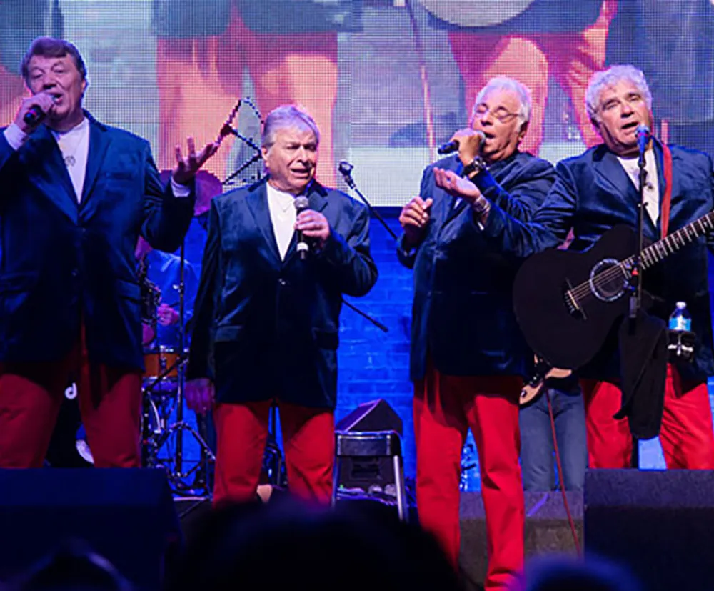 Four men are performing on stage likely singing while dressed in matching red trousers dark blazers and white shirts
