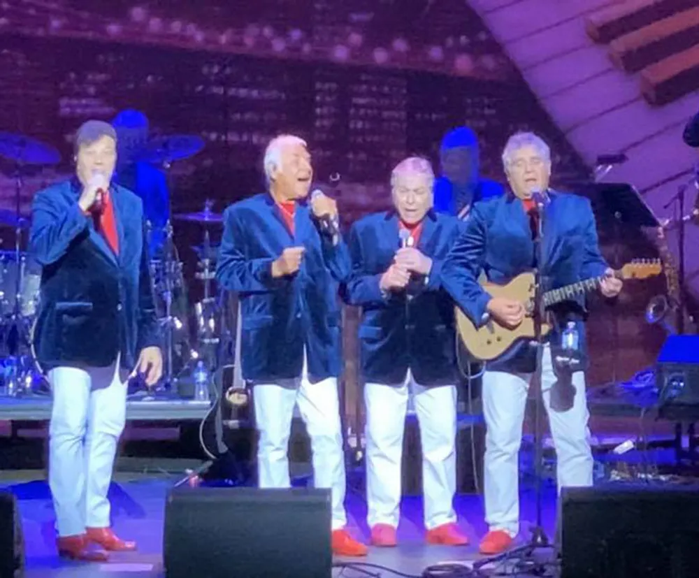 Four men in matching blue jackets red ties white pants and red shoes are performing onstage with three of them singing into microphones and one playing a guitar