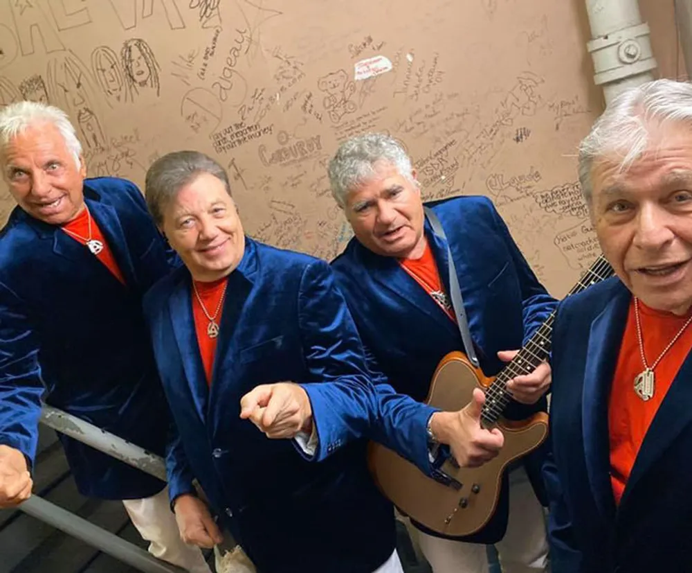 The image shows four men in matching blue blazers and red shirts one holding a guitar posing backstage with a wall filled with signatures and messages behind them