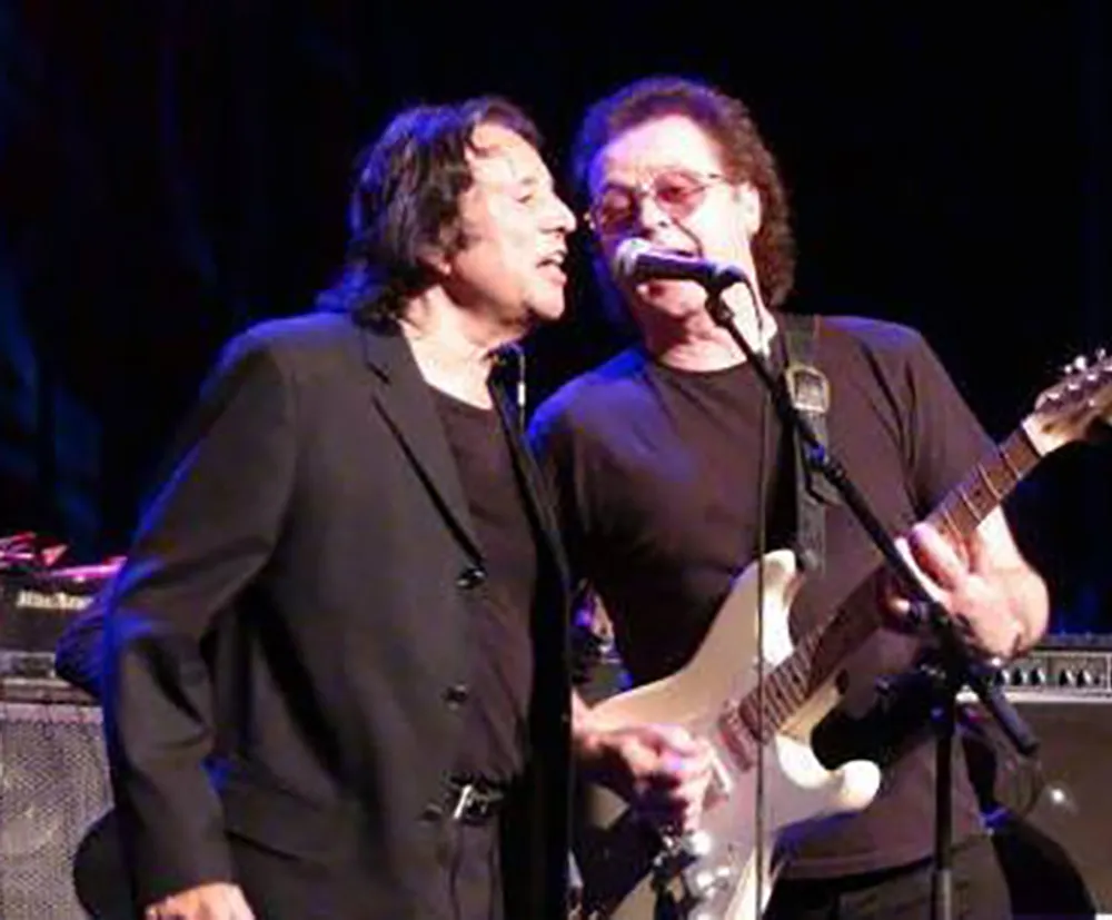 Two musicians are sharing a microphone on stage with one playing a guitar as they both sing into the same mic during a performance