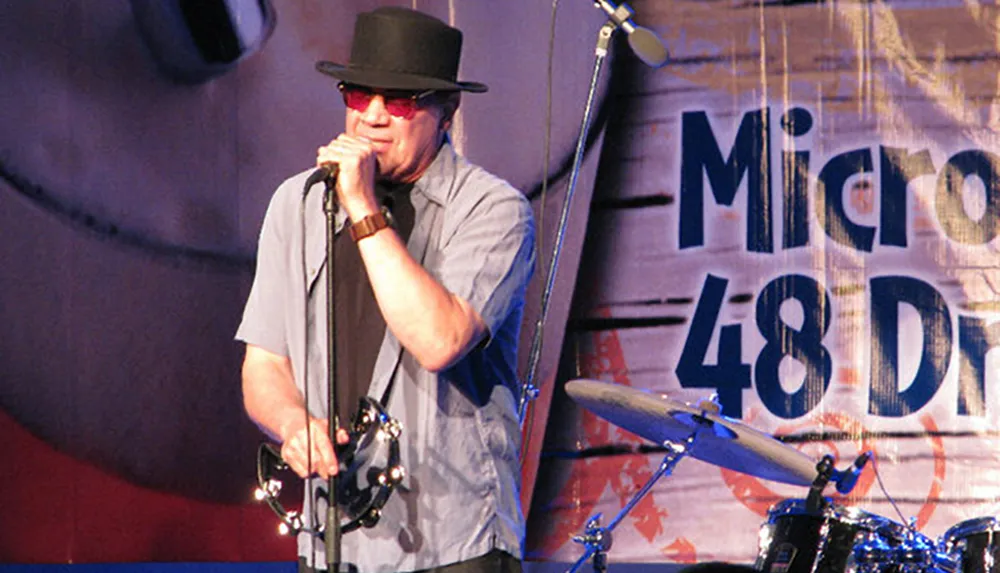 A musician wearing sunglasses and a hat performs into a microphone with musical equipment in the background